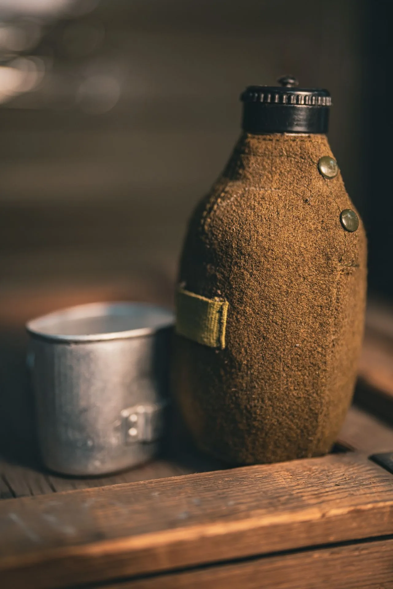 Romanian military canteen with olive wool felt cover, brass snap seam, and black bakelite screw cap, shown next to its nesting aluminum cup on a wood crate